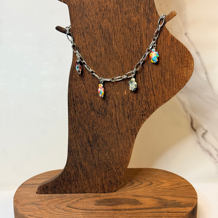 A silver-tone chain anklet with four colorful iridescent teardrop turtle charms displayed on a wooden neck stand.