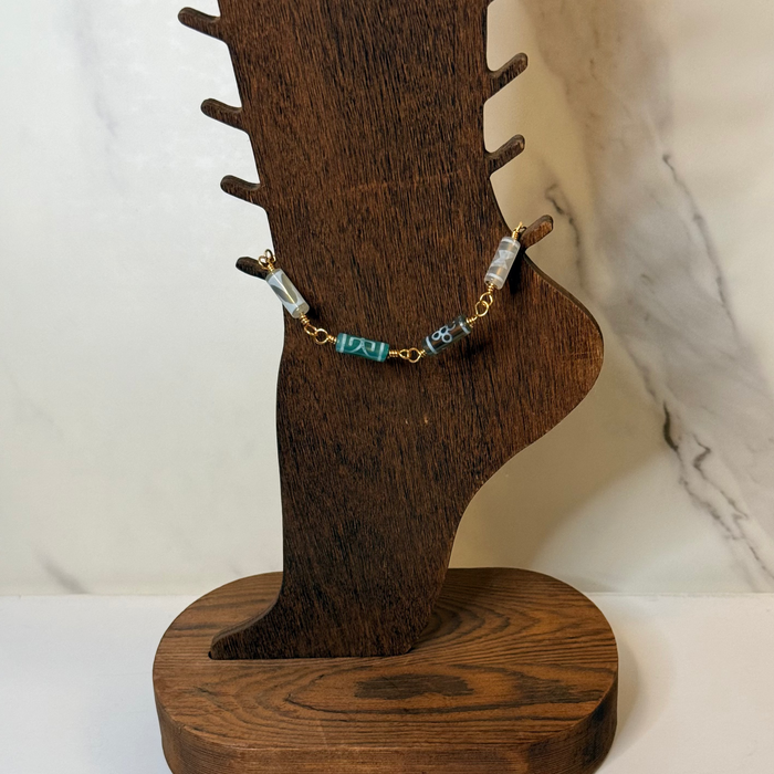 A gold chain anklet with green and white patterned beads displayed on a dark brown wooden jewelry stand.