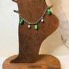 A silver chain bracelet with green and white bead charms displayed on a wooden stand.