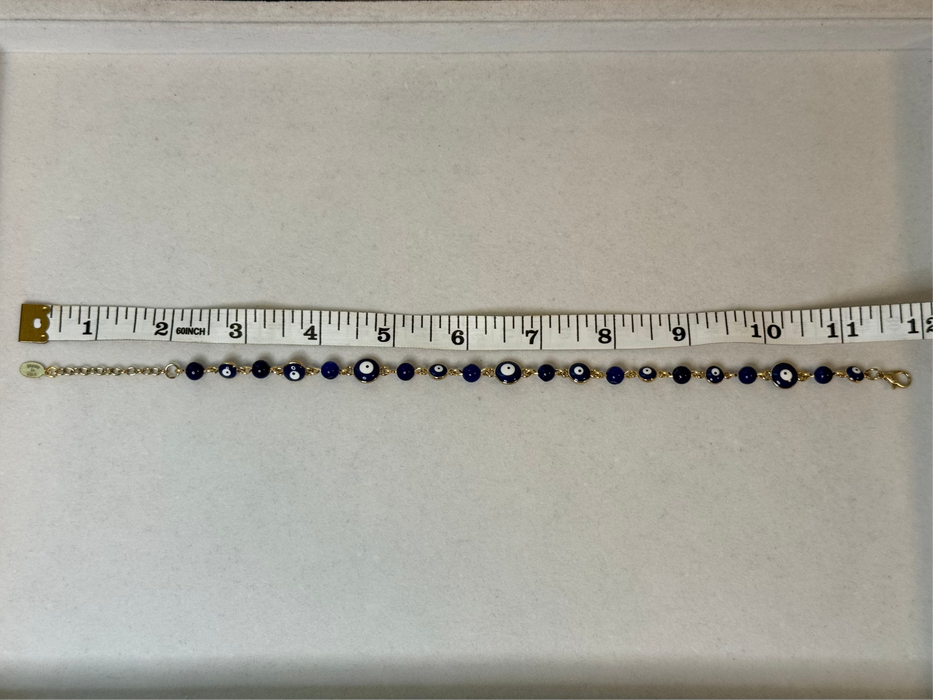 A gold chain bracelet with blue and white evil eye beads, shown on a beige surface with a measuring tape ruler above it for scale.