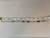 A gold chain anklet with multiple small cylindrical beads in green, gray, black, and white, shown on a light beige surface with a measuring tape ruler above it.