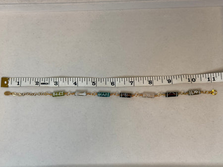 A gold chain anklet with multiple small cylindrical beads in green, gray, black, and white, shown on a light beige surface with a measuring tape ruler above it.