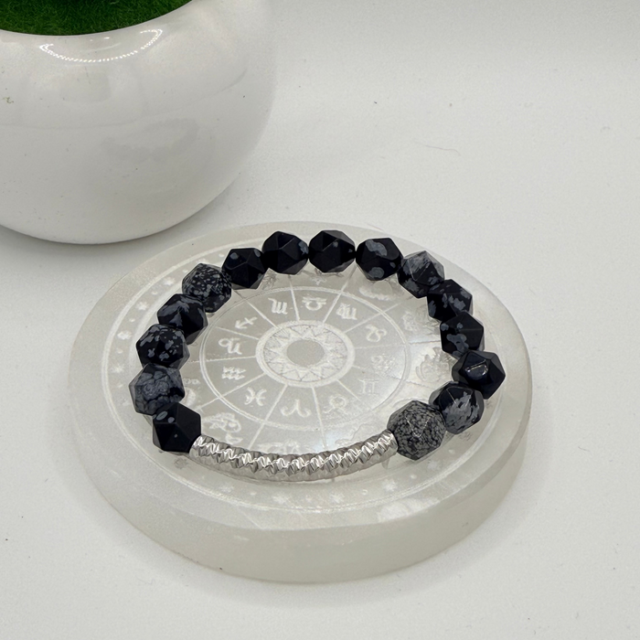 A beaded bracelet with faceted black and white gemstone beads and a silver-toned beaded accent section, displayed on a clear acrylic stand with a white background and a small potted succulent in the upper left.