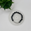 A beaded bracelet with black and white marbled beads and a silver-toned chain segment, displayed on a clear round stand next to a green potted plant on a white surface.