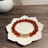 Reddish orange beaded bracelet with a gold clasp on a decorative white dish, next to a small plant.